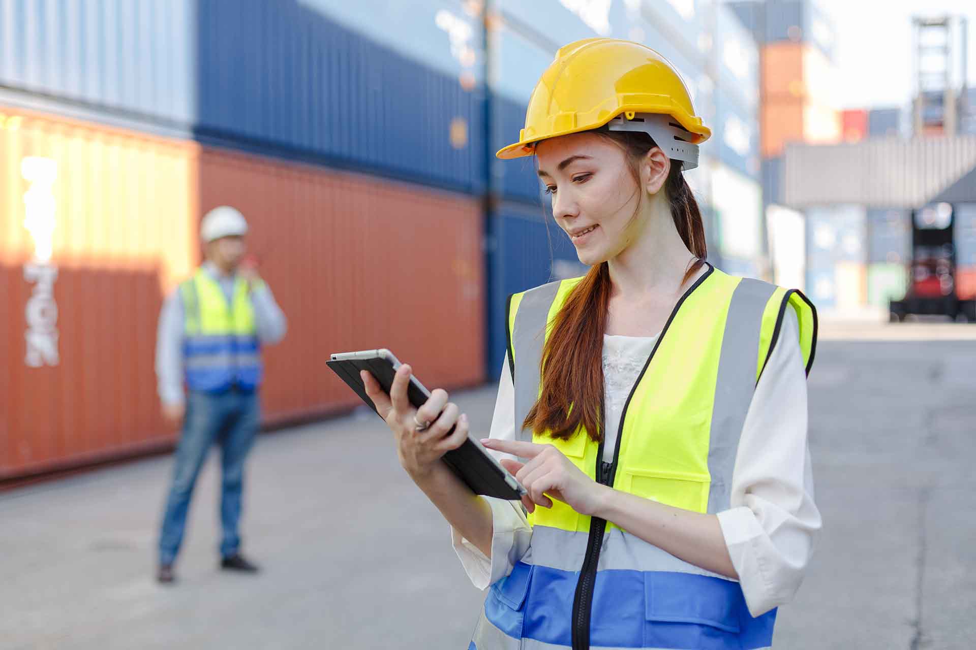 A woman wearing a hardhat and high visibility vest standing in front of shipping containers and holding a tablet.